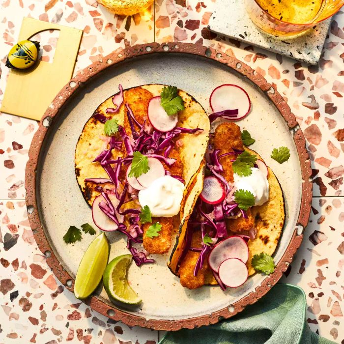 Two fish stick tacos topped with radishes and red cabbage, served on a round plate with lime wedges