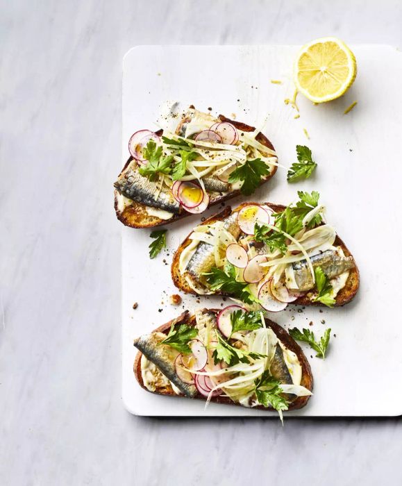 These Smoked Sardine Toasts are topped with lemon mayo and a crisp fennel salad, garnished with thinly sliced radishes, and served on a white cutting board with half a lemon.