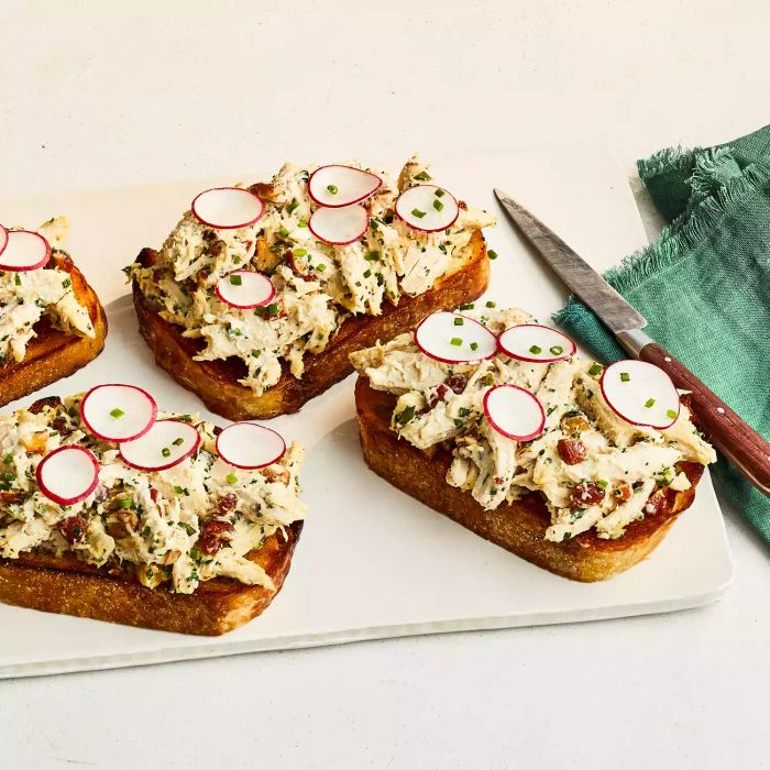 Four herby chicken salad tartines topped with radish slices, displayed on a white cutting board with a knife to the side