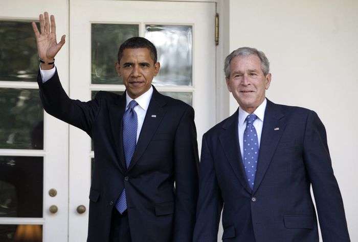 Two men dressed in suits stand together, one waving, outside a building with glass doors.