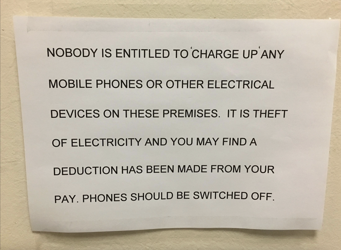Sign reads: "No one is permitted to 'charge' their mobile phones or any other electronic devices on these premises. This is considered theft of electricity, and pay deductions may apply. Phones must be turned off."