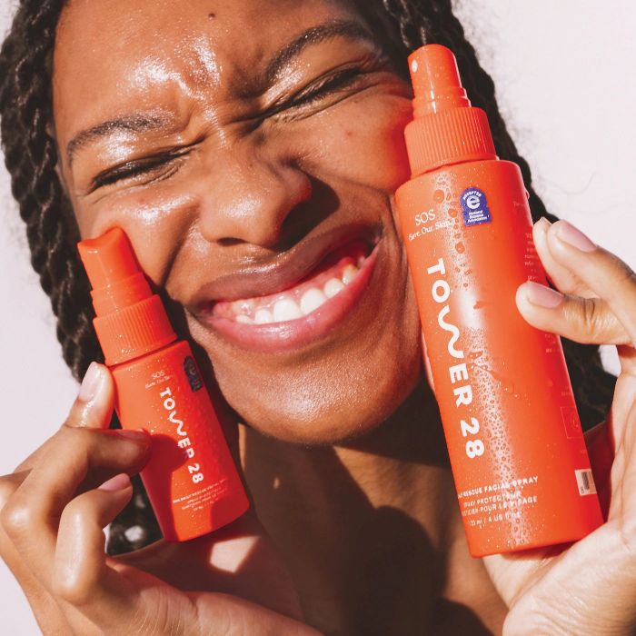 The model is holding two orange bottles of facial spray.
