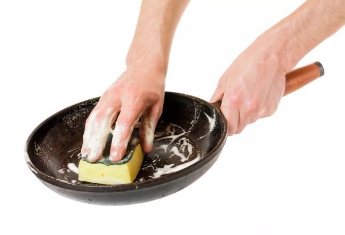 A close-up of a cast-iron skillet with dish soap and a sponge, set against a white background