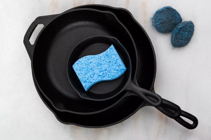 Cast-iron skillets accompanied by a blue sponge, displayed on a white background