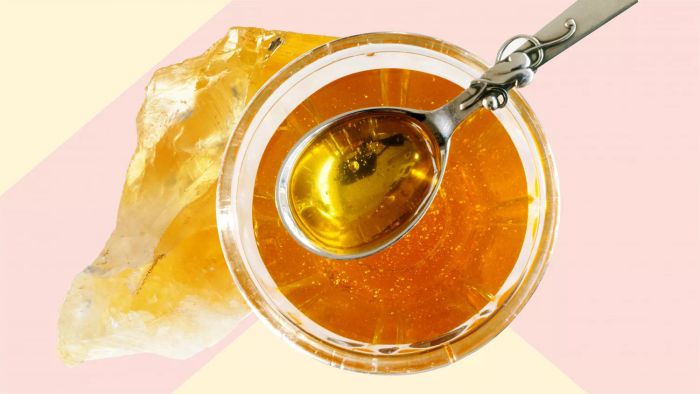 A glass bowl filled with honey, accompanied by a metal spoon beside crystallized honey