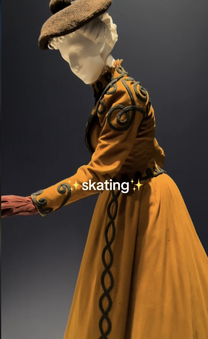 A mannequin sporting a vintage-style dress with elaborate patterns and a hat is posed as if skating. The word 