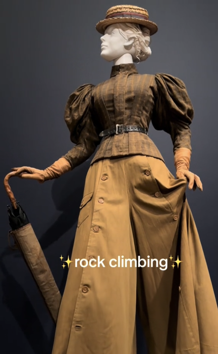 A mannequin dressed in a vintage feminine outfit, complete with a wide-brimmed hat and puffy sleeves, holds an umbrella. The text displayed reads, 