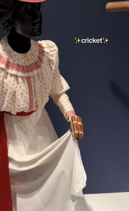 A display from the Victorian era shows a mannequin in a period dress and gloves, holding a cricket bat. The image features the text: 