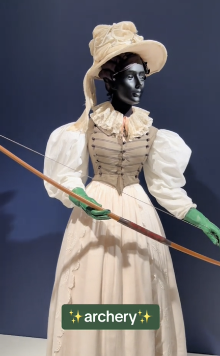 A mannequin in a vintage-inspired white dress, green gloves, and a large hat, holding a bow. Text reads 
