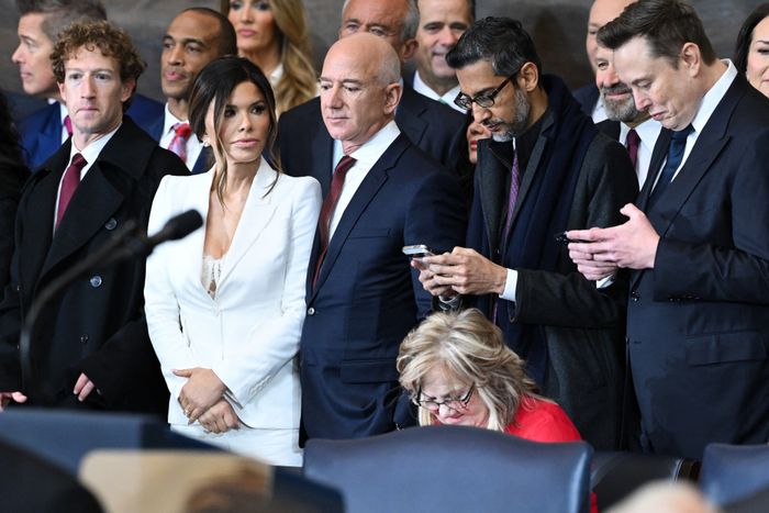 A group of people at a formal event, some in suits, with one woman wearing a white outfit. Several individuals are on their phones.