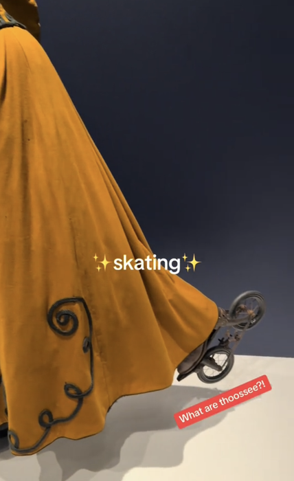 A person dressed in a long gown adorned with intricate designs is roller skating. The words 