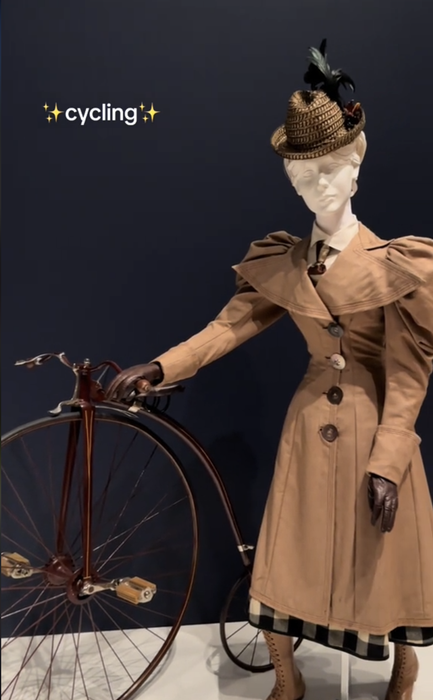 A mannequin in vintage cycling attire stands next to a large-wheeled bicycle. The caption at the top states, 