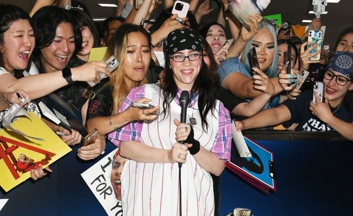 A person in casual clothing with a bandana, holding a microphone, surrounded by eager fans snapping photos and presenting items for autographs.