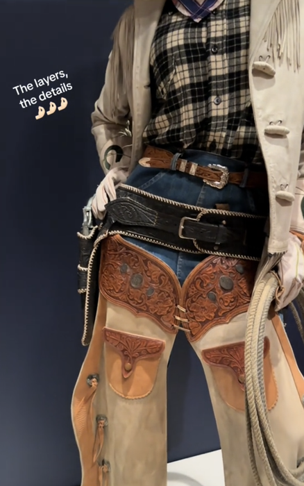 A close-up of a cowboy outfit displayed on a mannequin, featuring layers of clothing, decorative chaps, a plaid shirt, multiple belts, gloves, and a coiled rope. The caption reads: 