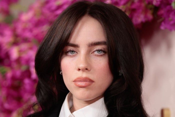 A person with long, wavy hair dressed in a formal collared shirt, standing in front of a vibrant floral backdrop.