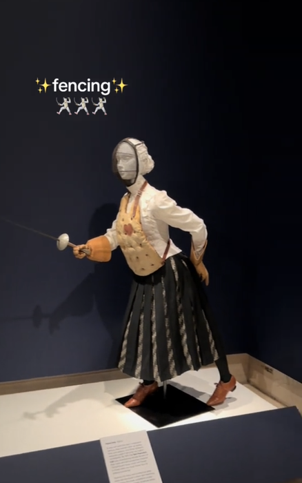 A historical museum display features a fencing outfit on a mannequin, complete with mask and sword, and the caption 