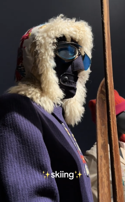 A person clad in a fur-lined hood, sporting ski goggles and holding vintage wooden skis, with the caption 