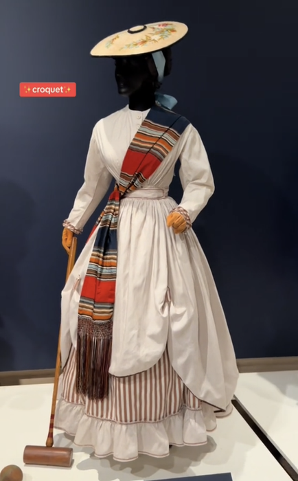 A mannequin dressed in a vintage Victorian-style croquet outfit, complete with a brimmed hat, striped sash, and holding a croquet mallet. The text 