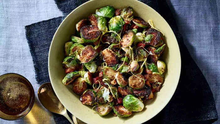 An overhead shot of roasted Brussels sprouts with pancetta served in a white bowl on top of blue fabric, accompanied by a metal spoon and a small metal bowl of peppered oil on the left side.
