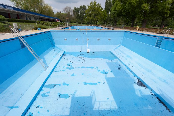 An abandoned outdoor swimming pool with ladders leading into it, its bottom covered in peeling paint, and surrounded by trees and buildings in the background.