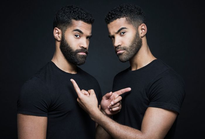 Two identical men in black t-shirts pointing at each other in front of a dark background.