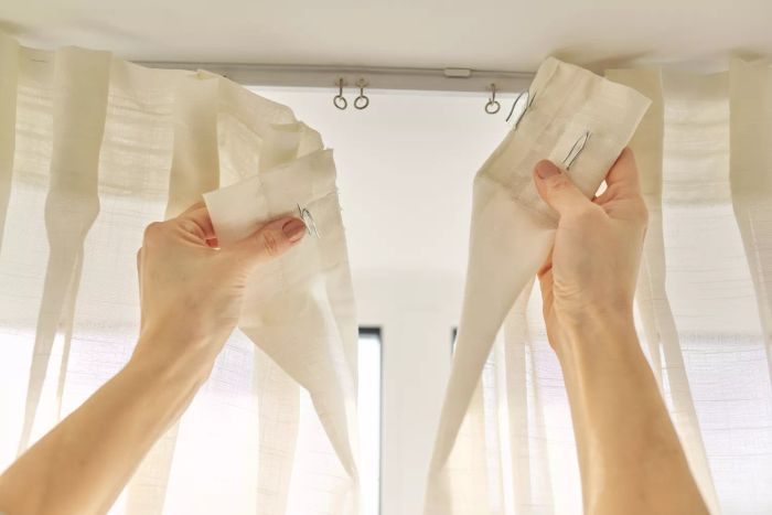 A close-up of a woman’s hands hanging curtains with metal hooks on the ceiling ledge.