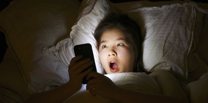 A person lying in bed, staring at their phone with a surprised expression.