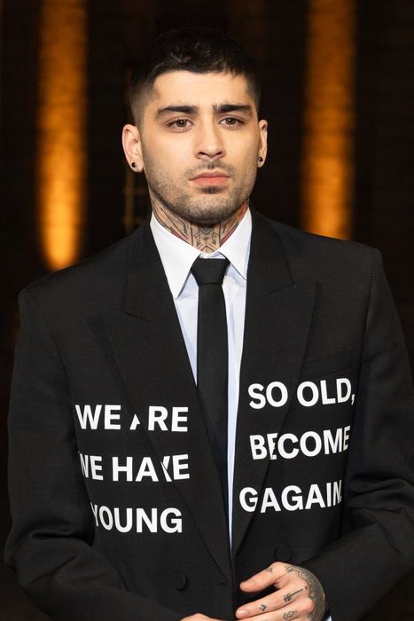 A person in a fashionable suit with a slogan, sporting facial tattoos, neatly styled hair, and rings, attending an event