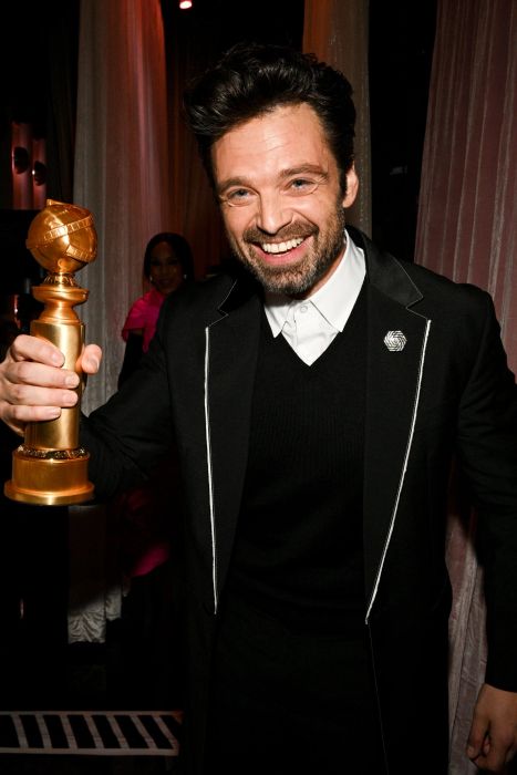 Sebastian Stan grinning while holding a trophy in a suit with a white shirt and black sweater at the award ceremony