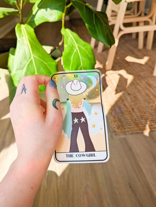 A tarot card-inspired window catcher featuring a blonde cowgirl with her back turned, wearing a white cowboy hat and jeans adorned with stars on the back pockets.