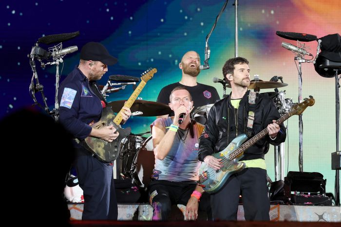 The band performs on stage with instruments; the singer kneels at the center, flanked by the guitarist and bassist. The drummer smiles in the background