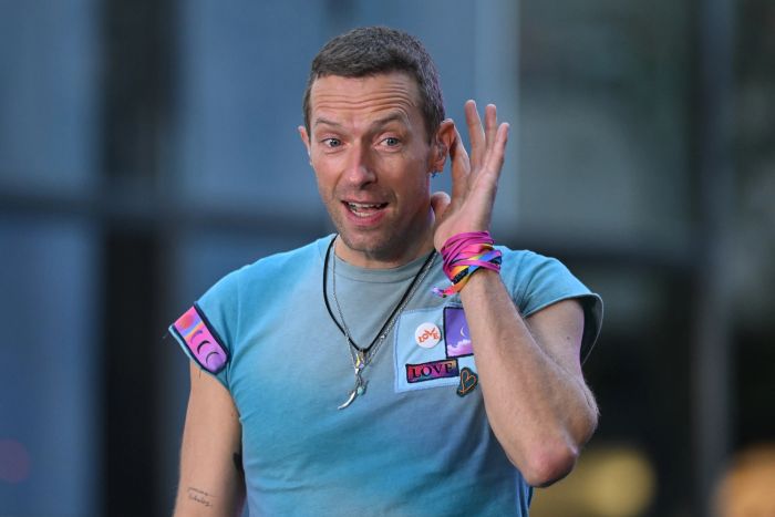 A person in a casual sleeveless shirt adorned with colorful patches and wristbands holds a hand to their ear, smiling in an outdoor setting