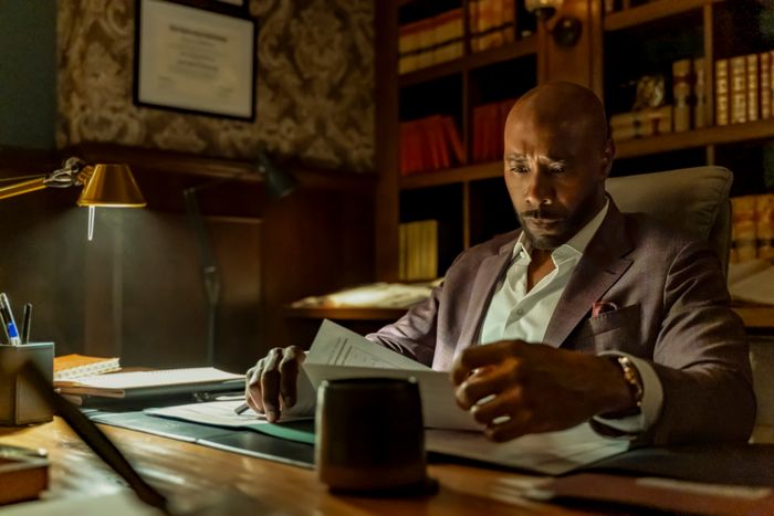 A man in a suit sits at a desk, absorbed in reading through documents, with a backdrop of bookshelves.