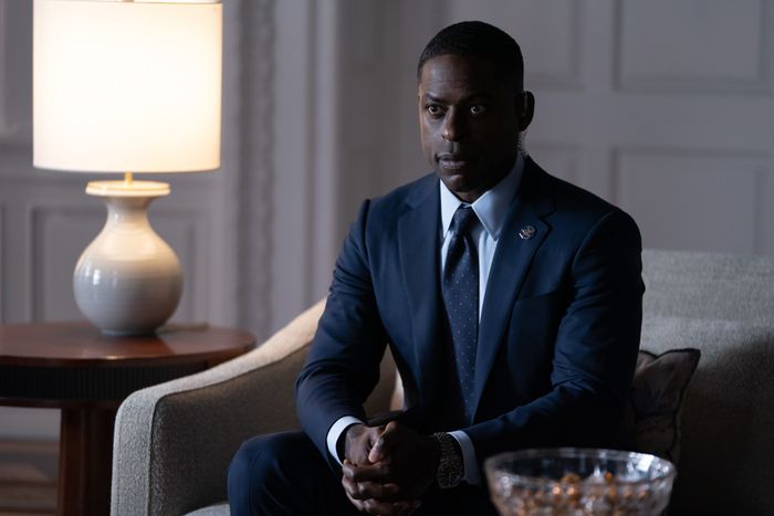 Sterling K. Brown dressed in a formal suit, sitting in a dim room, appearing pensive with a lamp beside him on a table