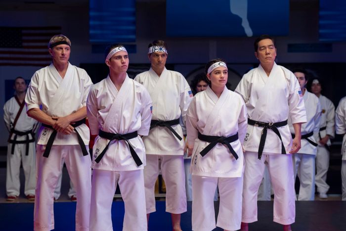 A group of five martial artists in karate gi, wearing black belts, stand in a focused and attentive pose within a dojo setting