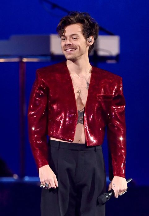 A person wearing a shimmering jacket and pants, holding a microphone while smiling on stage, with musical instruments in the background