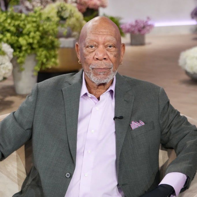 Morgan Freeman, dressed in a blazer and shirt, sits on a couch surrounded by potted plants, gazing at the camera.