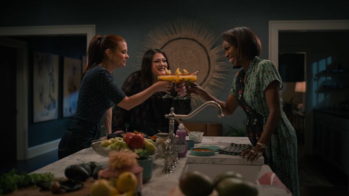 Three women, standing in a kitchen, raise their cocktail glasses in a cheerful toast, surrounded by fresh fruits and vegetables on the countertop.