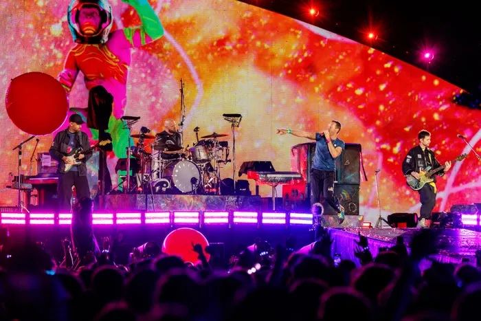 The band delivers an energetic performance on a vibrant stage, surrounded by a lively crowd, with colorful visuals displayed on a massive screen behind them