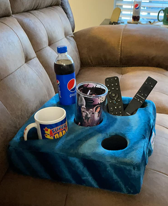 A blue pillow with five compartments, holding a can, a drink, a remote control, and a phone.