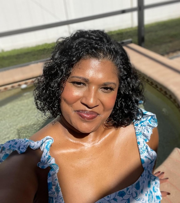 A selfie of a reviewer enjoying the sun by the pool, beaming with a smile, dressed in a floral-patterned sleeveless top.