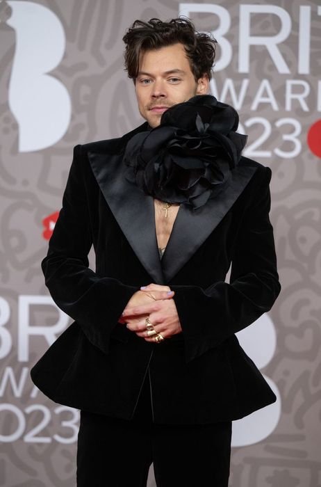 A person dressed in a black velvet suit with a large floral pin on the lapel, posing in front of a red carpet event backdrop