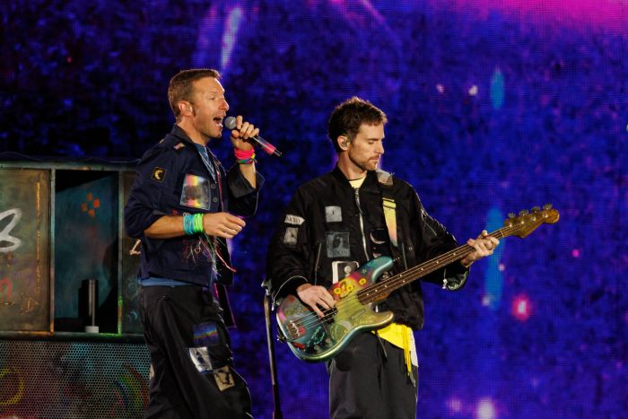 Two musicians passionately perform on stage. One belts out vocals into a microphone, while the other strums a guitar. Both sport casual, artistic jackets