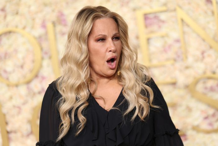Jennifer, with wavy hair and wearing a black ruffled top, makes an expressive face against a floral backdrop at a public event.