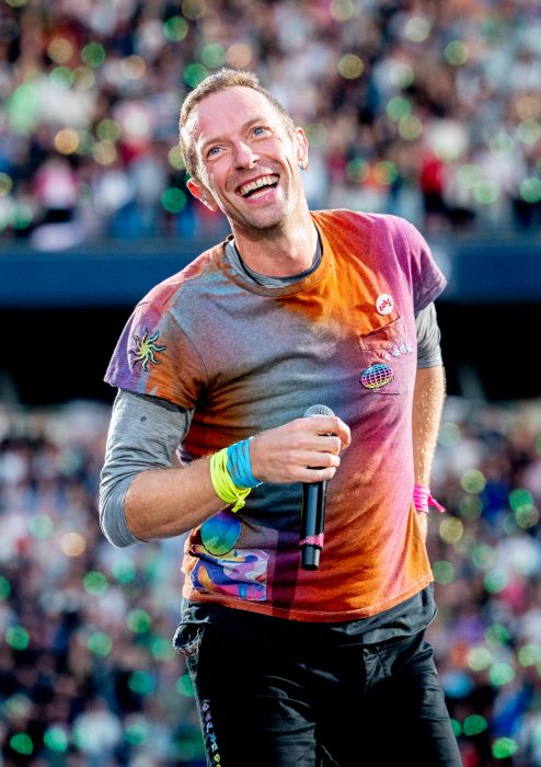 A person on stage beams with a smile, gripping a microphone while wearing a graphic t-shirt with a vibrant, colorful design. The crowd and bright stage lights set the atmosphere in the background.