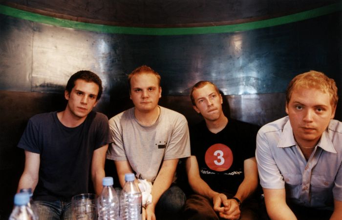 Four men sit close together, casually dressed, with water bottles placed on the table in front of them. The scene is set in a dimly lit room with a metallic backdrop