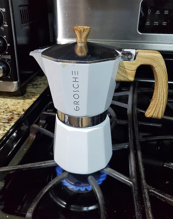 A white moka pot with a wooden handle brewing espresso on a lit stove.