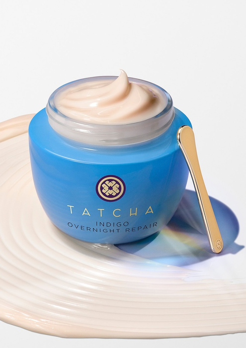 Tatcha Indigo Overnight Repair Cream displayed in an open blue jar with a delicate gold spatula resting beside it, placed on a textured surface