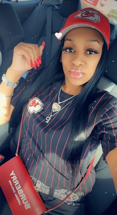 A reviewer dressed in a team logo sports jersey and cap, sitting in a car, with long hair, a cross necklace, and holding a small clutch bag.