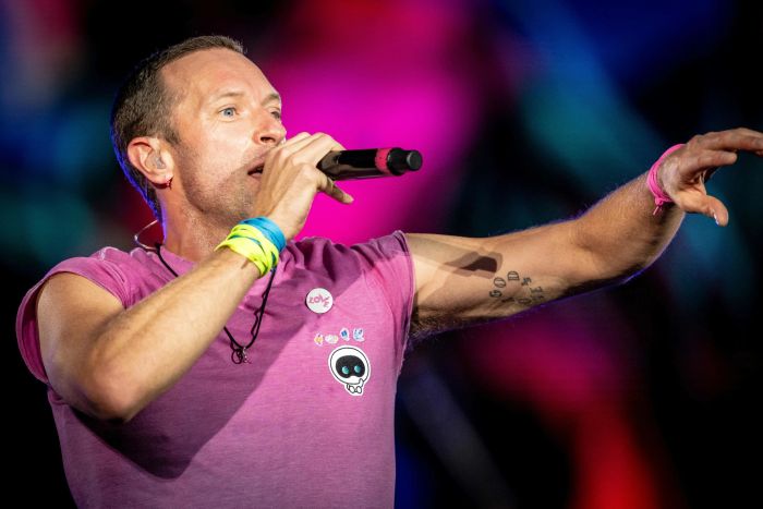 A person dressed in a short-sleeve shirt energetically performs on stage, gripping a microphone, with vibrant, colorful lights illuminating the background.